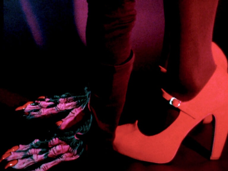 Detail against a dark background of a person wearing black tights and high heel orange shoes. They are wearing playful skeleton costume gloves with the shape of bones painted in pink, and reaching all the way down to the ground and the person's fingers are spread out on the ground beside their bright high heels