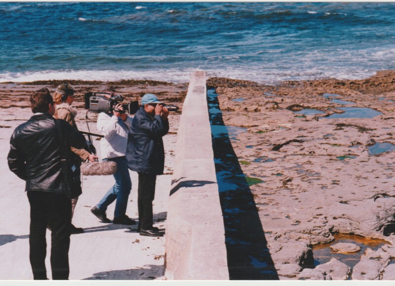 A low concrete wall spans from the rocky sea shore down into the sea. Four men working together are standing in a group together. They are looking over and beyond the wall, and are filming using two different cameras and audio equipment.