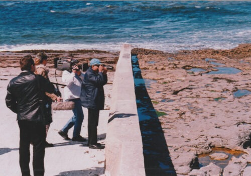 A low concrete wall spans from the rocky sea shore down into the blue sea. Four men working together are standing in a group. They are looking over and beyond the wall, and are filming using two different professional cameras and audio equipment