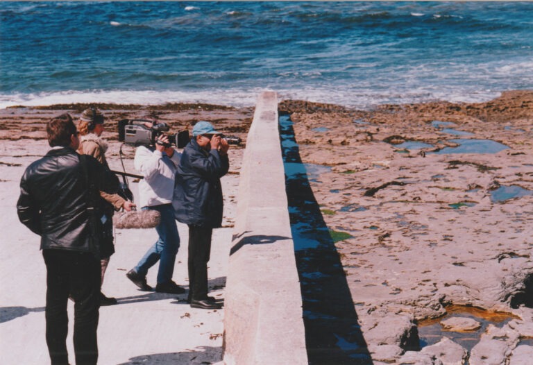 A low concrete wall spans from the rocky sea shore down into the blue sea. Four men working together are standing in a group. They are looking over and beyond the wall, and are filming using two different professional cameras and audio equipment