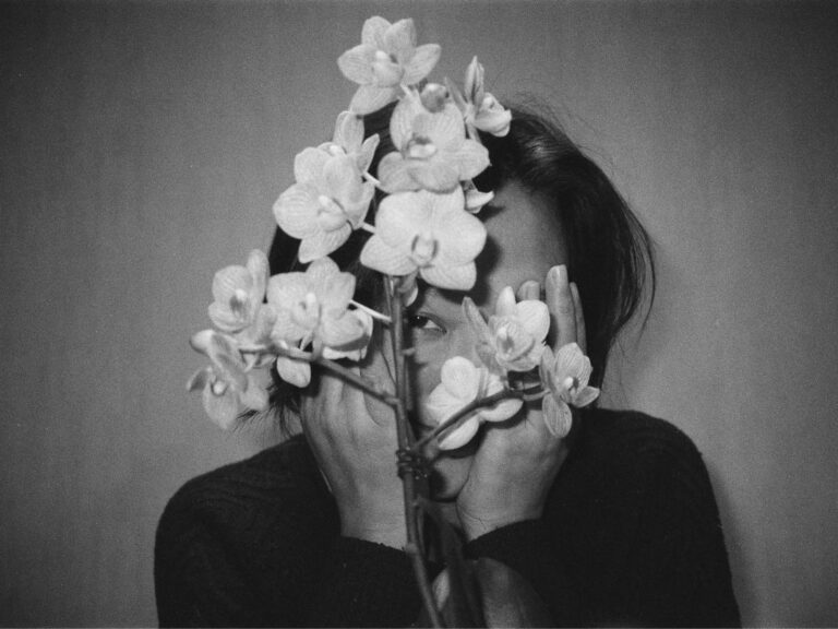 A black and white image of a person against a blank wall with flowers in front of their face, their hands are also raised in front of their face and one of their eyes peeks from in between the flower petals