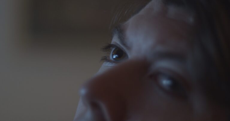 Still image from a film by Holly Márie Parnell. The image is a close-up of a white person's two eyes and nose. The room is dark but the person is slightly illuminated by the blue light from a computer out of frame.