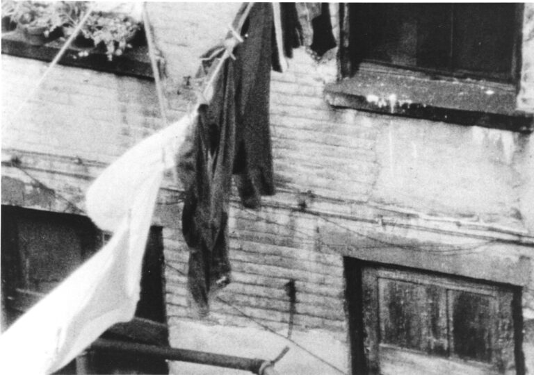 A still image from Betzy Bromberg's film Ciao Bella or Fuck Me Dead. A black and white image of clothes hanging on a line to dry, in between residential urban buildings over a street.