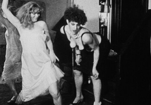 A still image from Tessa Hughes Freeland’s black and white film Baby Doll from 1982. Two young, slim women with voluminous hair are dancing together beside a floor-to-ceiling mirror. One woman has blonde, shoulder-length, wavy hair and wears a light slip dress. She is smiling and extends one arm back above her head while gleefully stepping forward with the opposite leg. The second woman has shorter, black curly hair, she is leaning forward and is moving her shoulders and arms rhythmically. She is wearing a black knee-length skirt, a black bra top that shows her cleavage, and black high heel sandals. Strings of white pearls are wrapped around her neck and one ankle