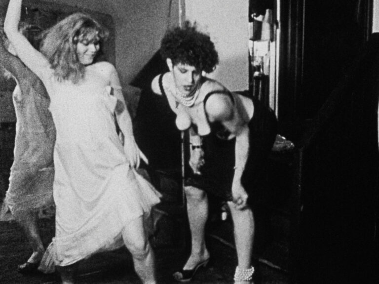 A still image from Tessa Hughes Freeland’s black and white film Baby Doll from 1982. Two young, slim women with voluminous hair are dancing together beside a floor-to-ceiling mirror. One woman has blonde, shoulder-length, wavy hair and wears a light slip dress. She is smiling and extends one arm back above her head while gleefully stepping forward with the opposite leg. The second woman has shorter, black curly hair, she is leaning forward and is moving her shoulders and arms rhythmically. She is wearing a black knee-length skirt, a black bra top that shows her cleavage, and black high heel sandals. Strings of white pearls are wrapped around her neck and one ankle