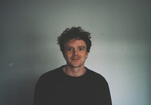 A colour headshot of Frank Sweeney, one of the film artists who will speak at aemi's upcoming online event 'Building a Practice in Artist Film'. Frank is a white man with short curly brown hair, wearing a black jumper. He is smiling and is sitting in front of a blank grey wall, light shines in from one side. The photograph is a polaroid and a white border frames the image.