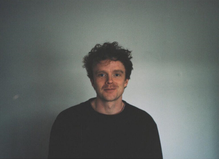 A colour headshot of Frank Sweeney, one of the film artists who will speak at aemi's upcoming online event 'Building a Practice in Artist Film'. Frank is a white man with short curly brown hair, wearing a black jumper. He is smiling and is sitting in front of a blank grey wall, light shines in from one side. The photograph is a polaroid and a white border frames the image.