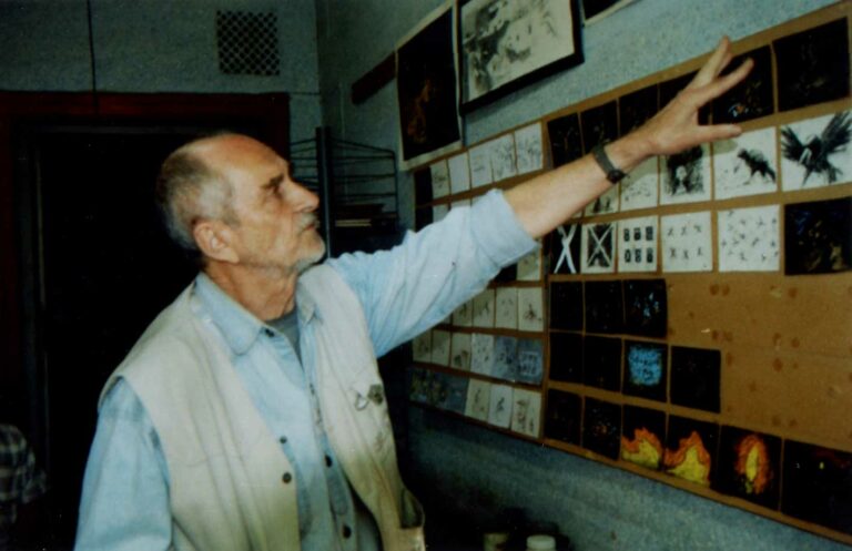 A man with pale skin stands in his art studio and points at a number of drawings on the wall