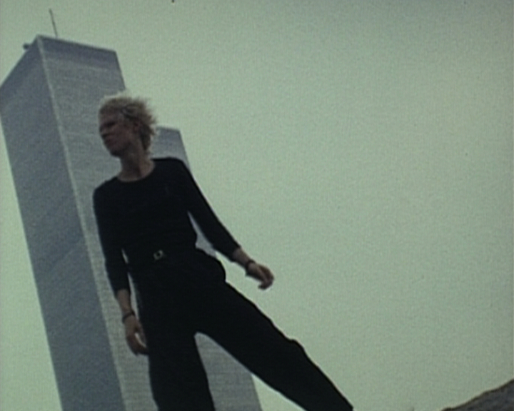 A still image from Vivienne Dick’s 1978 film She Had Her Gun All Ready. A woman with light skin and cropped blonde hair stands tall against the backdrop of a skyscraper in the distance. The woman’s clothing of black trousers and a black long sleeve top is stark against the minimalist background. The 16mm footage is taken from below at an angle, and the woman’s wide-legged stance creates further angular shapes within the composition of the image.