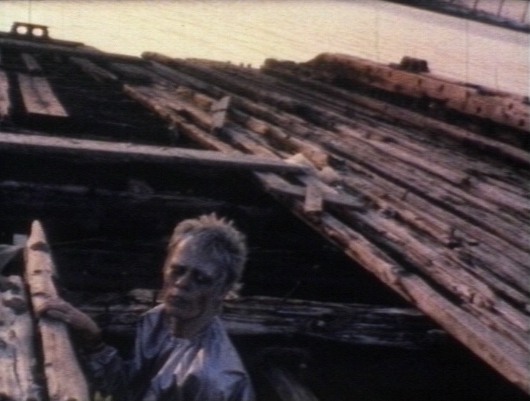 A still image from Vivienne Dick’s 1978 film called Staten Island. A person emerges from between broken wooden planks at a dockyard. They have light skin and their face is painted silver overall, with black under their eyes and dark red streaks on their cheeks. The cropped spiky blonde hair, and wearing a metallic silver suit, Warm daylight bounces off the water in the background.