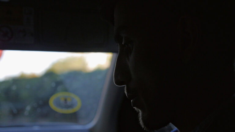 A still image from the digital film I Must Away by Dennis Harvey. The camera focuses on the profile of a young man's face, who sits inside a vehicle in darkness. Details of his face cannot be seen in the darkness but the light catches on his short facial hair and his eye gaze is directed downwards.