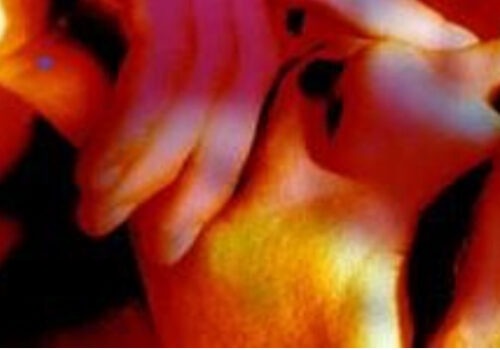 A still image from George Barber's video artwork called Ansaphone from 1996. The image is a close-up of a person's face with their hand held up to their mouth. Strong light in yellow, pink and orange colours shines on the person. The footage is shot at an angle from above the person's head, causing dark shadows around their eyes.