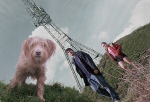 At a dynamic angle, a camera points up from the ground at a beige scruffy dog, and a teenage girl and boy. behind them is a grassy bank and a tall pylon