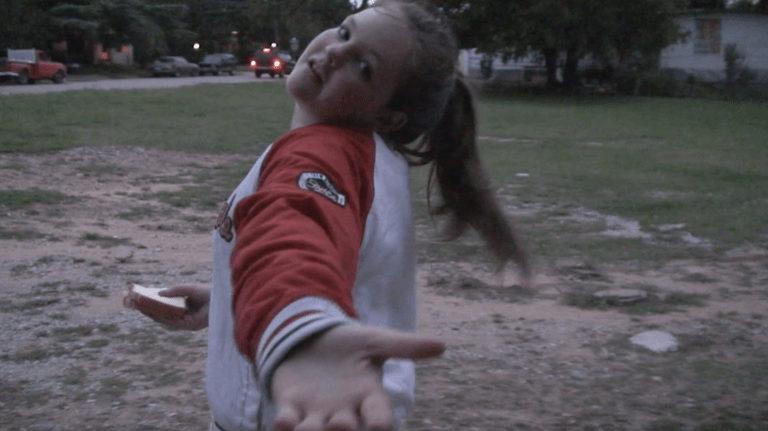 A still image from a film of a young girl with pale skin wearing a sports jacket and posing to the camera with her hand reaching towards the viewer