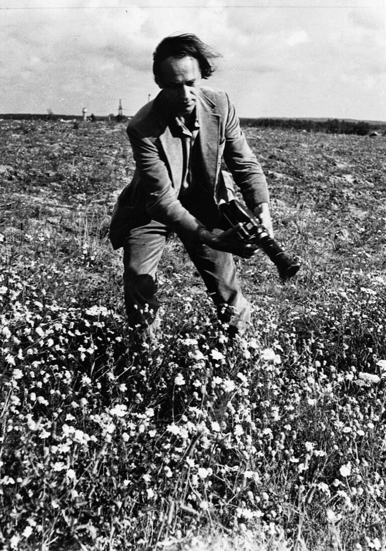 A black and white portrait photograph of man with pale skin in the middle of a field of flowers. There is wind flowing through his balding hair and he is animatedly carrying a black analogue camera by his side, his arms swinging as he points the lens down toward the mass of flowers.
