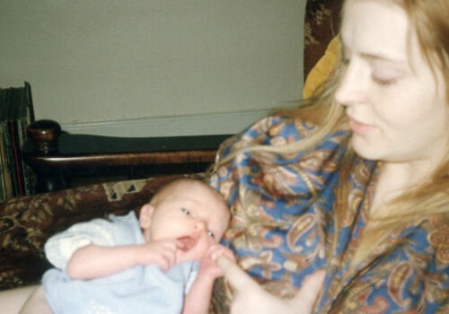 A still image from Myrid Carten's film called Sorrow had a baby. A young, white woman with long blonde hair sits in a chair and looks down at the white baby in her arms. The newborn baby is clutching the woman's finger and pulling it toward its open mouth. The woman is wearing a paisley print top in blue and brown colours, and a row of vinyl records is stored behind them.