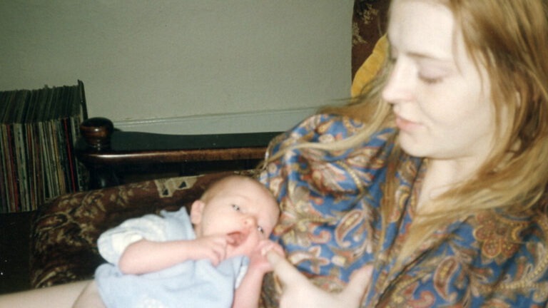 A still image from Myrid Carten's film called Sorrow had a baby. A young, white woman with long blonde hair sits in a chair and looks down at the white baby in her arms. The newborn baby is clutching the woman's finger and pulling it toward its open mouth. The woman is wearing a paisley print top in blue and brown colours, and a row of vinyl records is stored behind them.