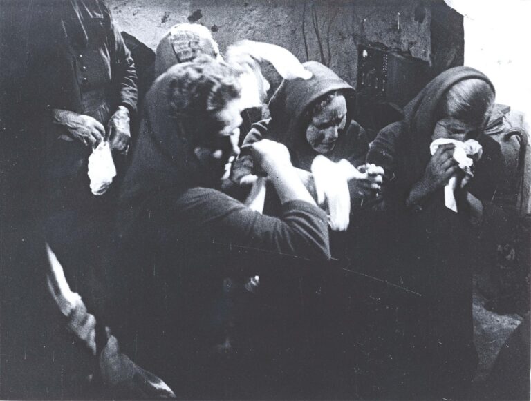 A still image from a black and white film of a group of women wearing dark traditional dresses and head shawls crying into white handkerchiefs