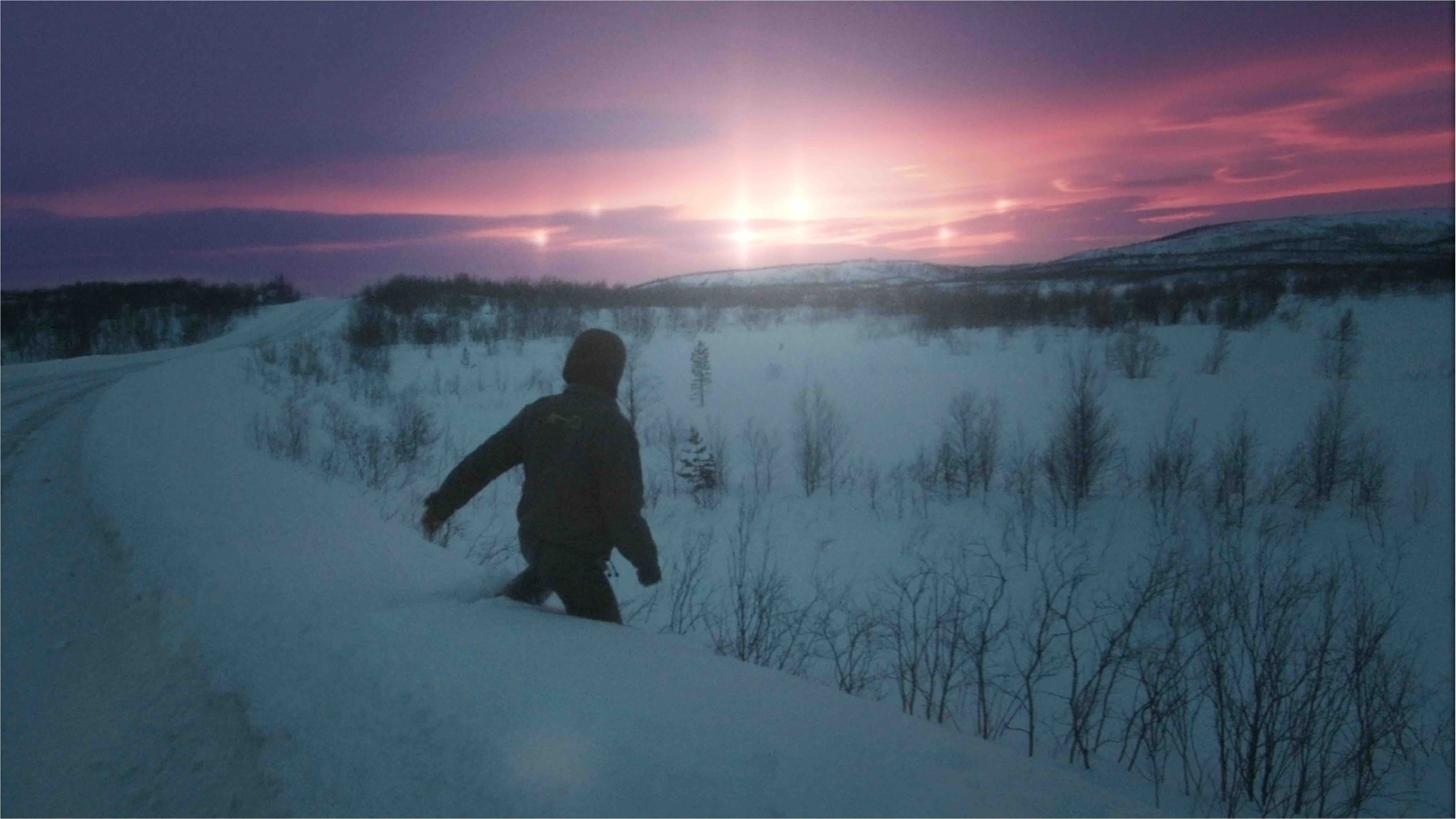 A snowy landscape scene featuring a person walking through deep snow down a slope where bare bushes peek through the snow. The sun is over the horizon, peeking through the clouds in a vivid purple and pink sky.