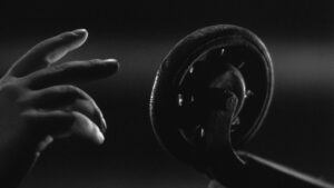 Still from Charlie Shackleton's 35mm film called The Afterlight. In a very dark black and white close-up shot, soft light highlights the outline of an adult's hand reaching towards an old domestic wheel