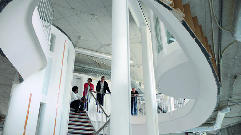 A still image from Wendelien van Oldenborgh’s digital video artwork Prologue: Squat/Anti-Squat from 2016. A group of at least four adults of diverse ethnicities are gathered at the top of a staircase inside a large building, conversing with one another. The angle of the camera highlights the building’s architectural features, including curved internal balconies, pillars painted white, a semi-circular room overlooking the staircase, and the concrete ceiling. The scene is shot in the daytime and the image is bright, with daylight bouncing off the white surfaces. Some inner-workings of the building, such as pipes and structural supports, are exposed under one ceiling.