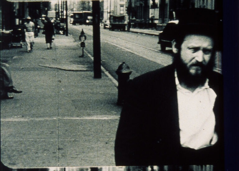 A black and white still image of a Jewish man with pale skin walking down the road