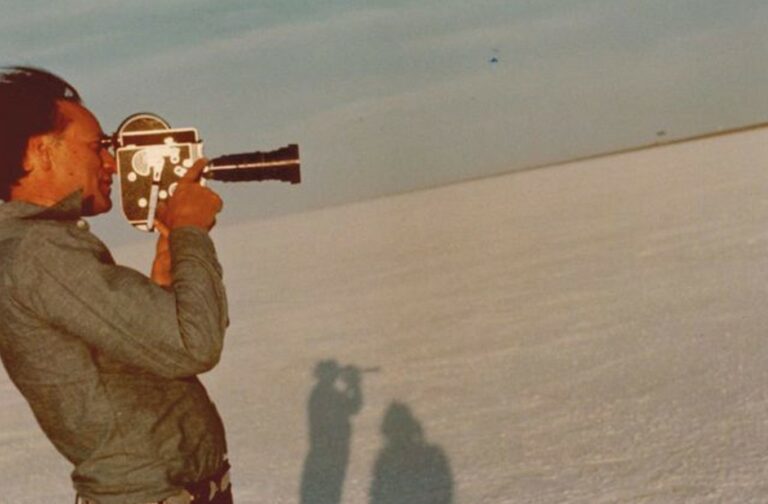 On a bright sunny day, a man with pale skin and dark hair holds a hand-held analogue camera to his eye and points it towards a vast empty landscape