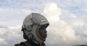 A child with dark skin wearing a futuristic helmet, stands outdoors with trees in the background, closes their eyes and directs their face towards the sky
