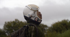A child with dark skin wearing a futuristic helmet, stands outdoors with trees in the background, closes their eyes and directs their face towards the sky