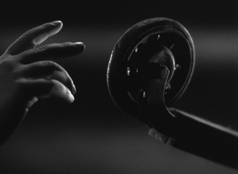 A very dark black and white still image from a film. In a close-up shot, soft light highlights the outline of an adult's hand reaching towards an old domestic wheel