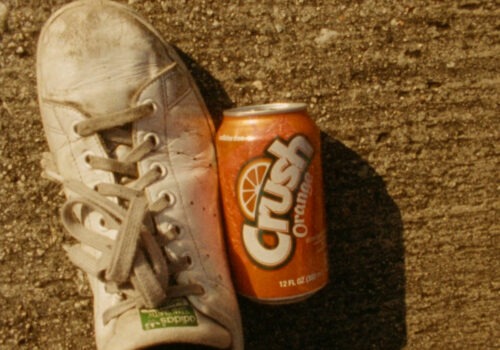An organge can of a fizzy drink called Crush on the ground beside a person's foot wearing a grubby white running shoe