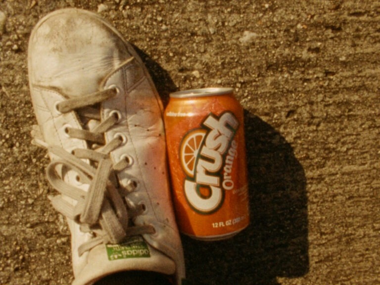 An organge can of a fizzy drink called Crush on the ground beside a person's foot wearing a grubby white running shoe