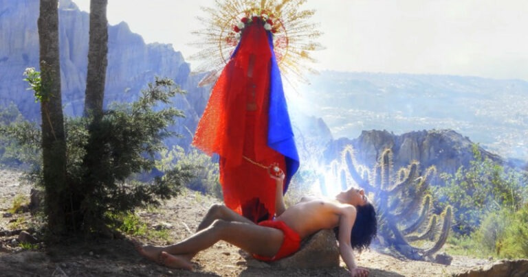 A person dressed similarly to the religious Madonna figure with a gold halo stands over a reclining figure lying on a rock in a dry, sunny landscape