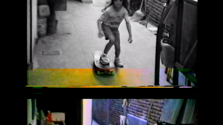 A still from an analogue video of a child skateboarding through a corridor space with stuff on either side. The child has long hair and is wearing tshirt and shorts