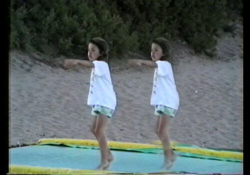 A still from an analogue video of a child jumping on a beach. The child is wearing a tshirt and shorts and their image is doubled so they appear twice in repetition