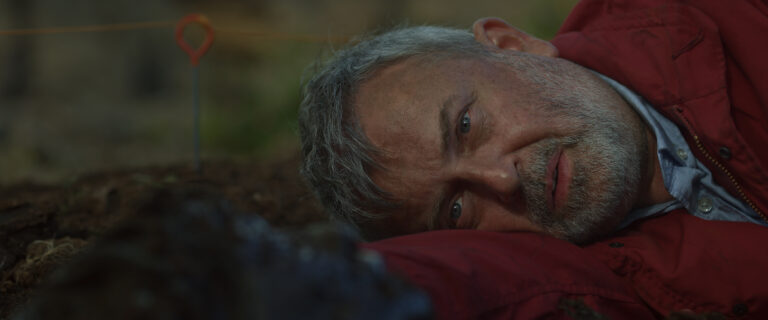 A close up of a male archaeologist with light skin lying on the ground during a dig in the bog