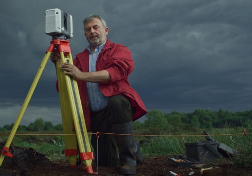 A male archaeologist with light skin kneels at his tripod and tools onsite during a dig in the bog