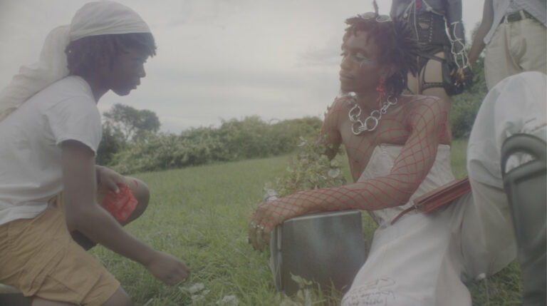 A washed out image taken in green landscape of a Black child reaching out to a Black queer person who is wearing red fishnet sleeves and a cream corset and trousers
