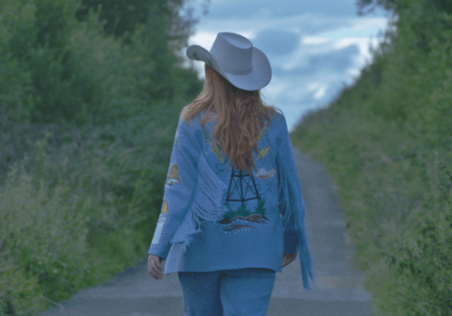 This is a still from the film '2 Channel Land' (2022) by Frank Sweeney. A person with strawberry blonde hair walks along an empty road that marks the border between two areas in Northern Ireland. The road's surface shifts from one shade of grey to another, clearly indicating the presence of a border line. It is a country lane is flanked by tall green bushes and trees on either side. The camera is positioned behind them, and there is a cloudy, grey-blue sky ahead. They are dressed in a white Stetson hat, sky-blue trousers, and an embellished sky-blue jacket with blue fringe on the sleeves. The jacket features an image of a radio transmitter on the back, surrounded by lightning bolts, a line drawing of the Irish border, clouds, grass, and soil.