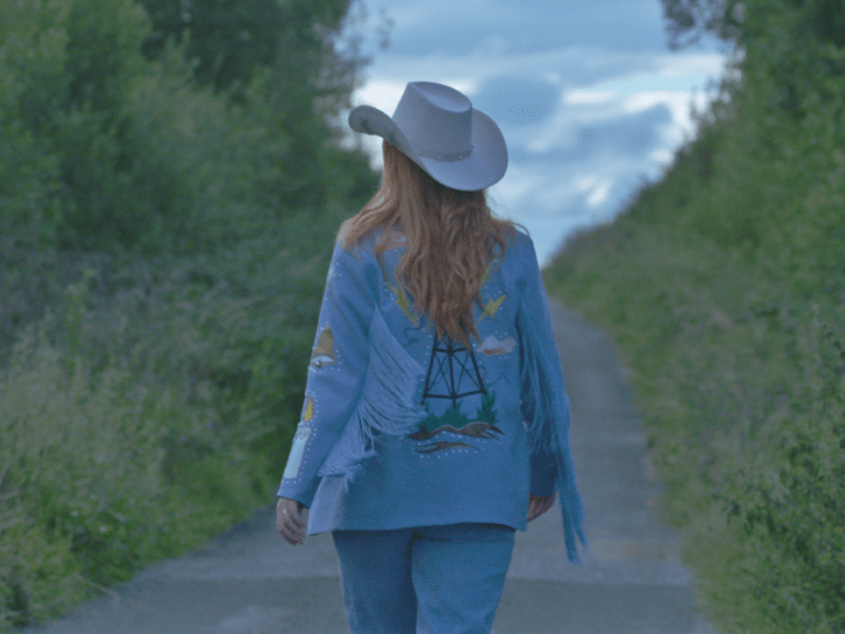 This is a still from the film '2 Channel Land' (2022) by Frank Sweeney. A person with strawberry blonde hair walks along an empty road that marks the border between two areas in Northern Ireland. The road's surface shifts from one shade of grey to another, clearly indicating the presence of a border line. It is a country lane is flanked by tall green bushes and trees on either side. The camera is positioned behind them, and there is a cloudy, grey-blue sky ahead. They are dressed in a white Stetson hat, sky-blue trousers, and an embellished sky-blue jacket with blue fringe on the sleeves. The jacket features an image of a radio transmitter on the back, surrounded by lightning bolts, a line drawing of the Irish border, clouds, grass, and soil.