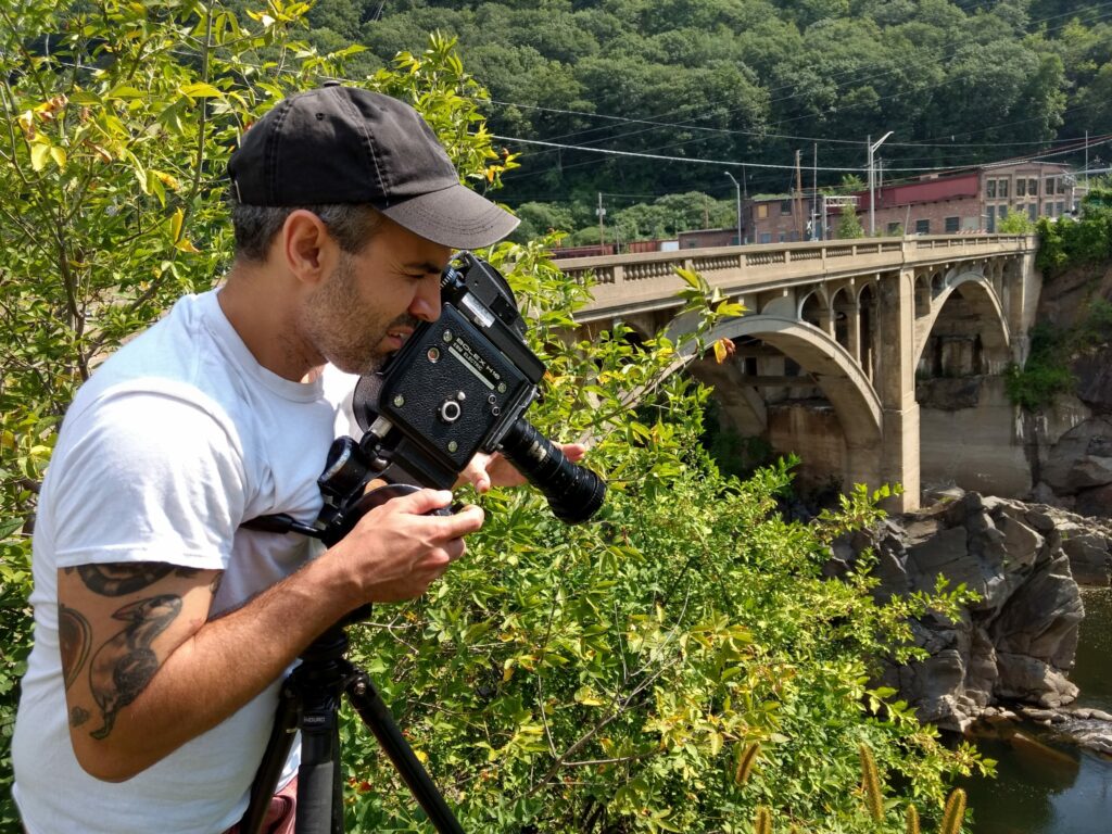 man with animal tattoos wearing a white t-shirt and a baseball cap looks into the lens of his bolex camera while shooting from a height. He's surrounded by green bushes and a stone arch bridge is in the background