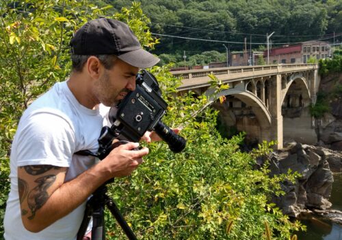man with animal tattoos wearing a white t-shirt and a baseball cap looks into the lens of his bolex camera while shooting from a height. He's surrounded by green bushes and a stone arch bridge is in the background
