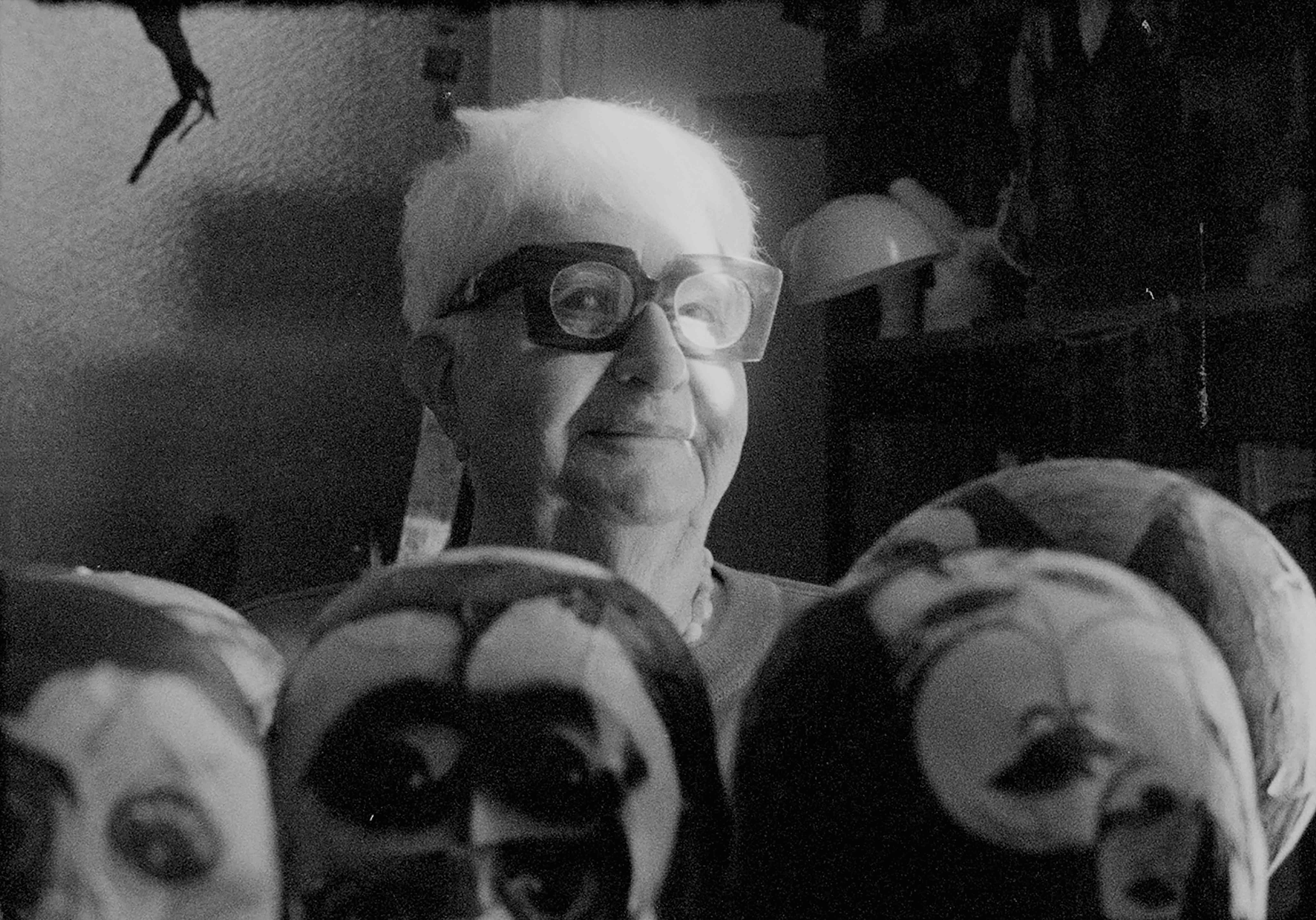 Black and white image of a woman with cropped grey hair and large stylish glasses smiling and sitting amongst life-size sculptural heads in the foreground