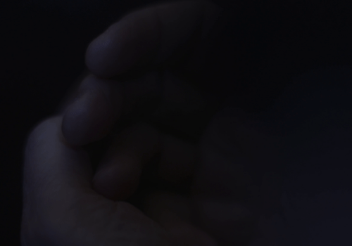 A dark close-up image of a person's hands loosely clasping each other