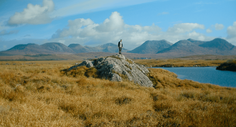 A still image from a film of a landscape. In the distance is blue sky, puffy clouds and a mountain range. Surrounded by a natural terrain of long brown grass, in the centre of the image a person stands on top of a large rock and looks toward a large area of blue water
