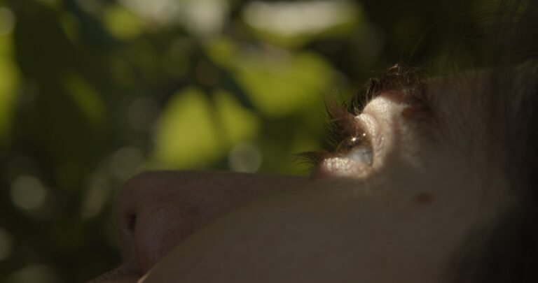 This is a still from the short film 'Cabbage' (2023) by Irish filmmaker Holly Márie Parnell. This image features a side angle view of a man's face with light skin and brown hair in a lightly shadowed space. A bright spot of daylight illuminates his left eye, and we can also see part of his cheek and nose in the frame in the light shadow as he looks upwards. The background displays out-of-focus green foliage and dappled light.