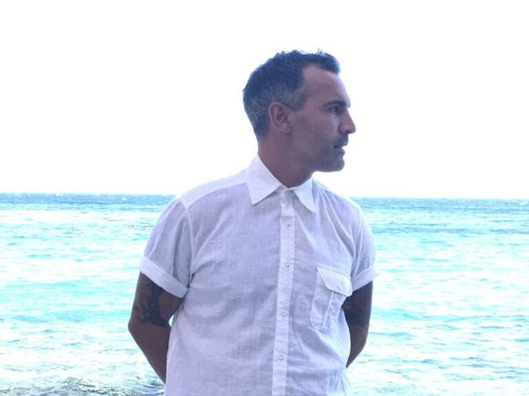 A man with tanned light skin wearing a white shirt stands with his hands behind his back in front of the bright sea