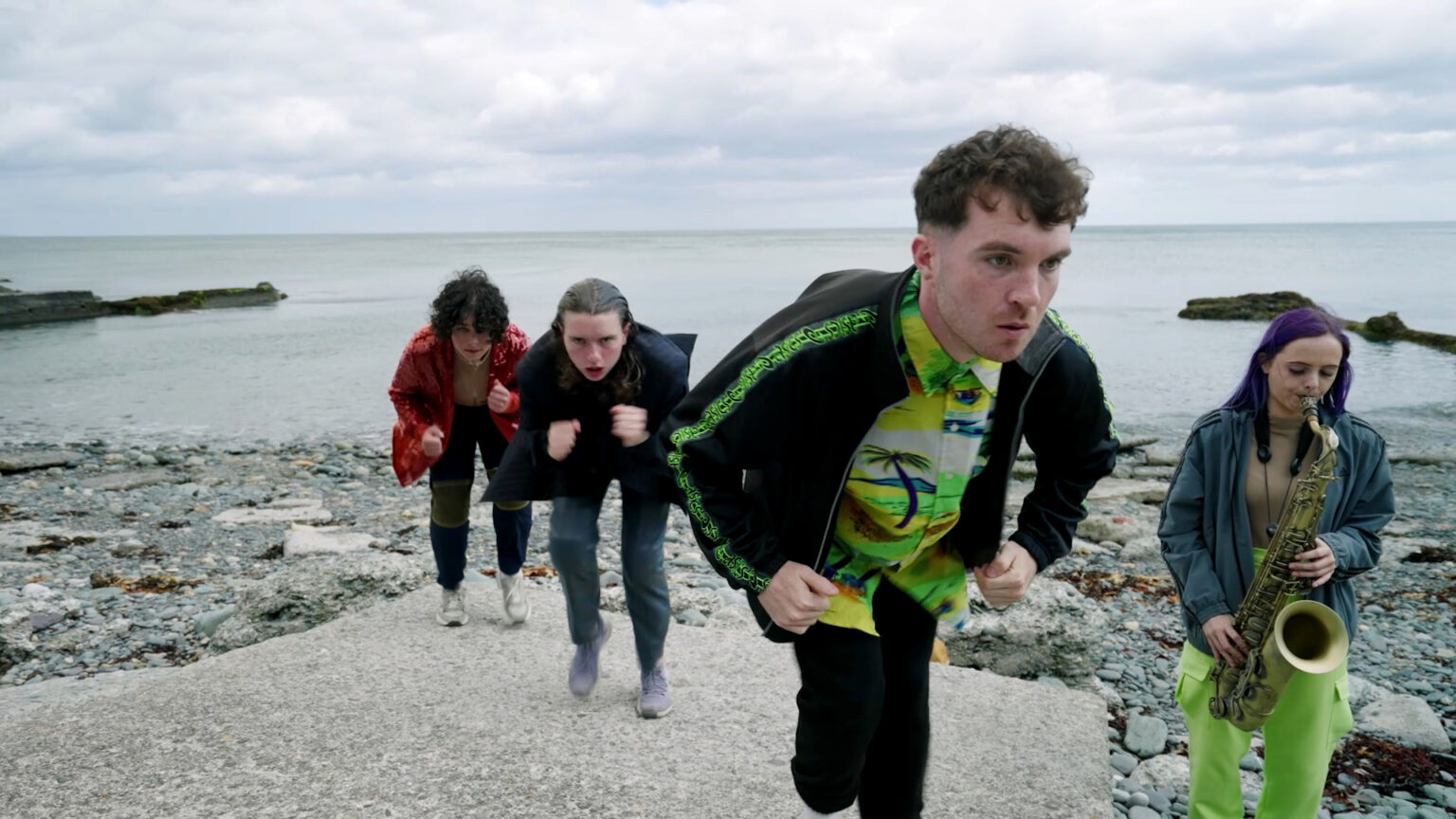 Still from video of a performance on a stony beach. Four young adults with light skin wear a mix of dark and bright clothing, including: a red sparkly jacket, lime green khaki pants, a yellow and a green Hawaii shirt under a casual black zipper with a green stripe down the sleeve that repeats the ASICS logo. The three people in the centre of the image are on a protruding rectangle of concrete. They have concentrated looks on their faces, and their bodies mirror each other while they hunch down to hold a "run on the spot" position. The person on the right hand side has purple hair and is playing the saxophone. Behind them the sea is grey and the sky is totally overcast.