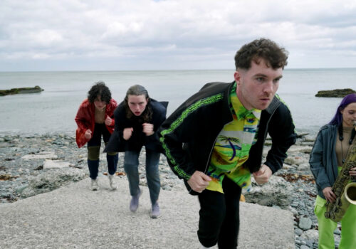 Still from video of a performance on a stony beach. Four young adults with light skin wear a mix of dark and bright clothing, including: a red sparkly jacket, lime green khaki pants, a yellow and a green Hawaii shirt under a casual black zipper with a green stripe down the sleeve that repeats the ASICS logo. The three people in the centre of the image are on a protruding rectangle of concrete. They have concentrated looks on their faces, and their bodies mirror each other while they hunch down to hold a "run on the spot" position. The person on the right hand side has purple hair and is playing the saxophone. Behind them the sea is grey and the sky is totally overcast.