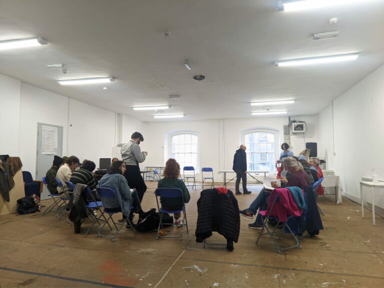 A group of students sit in a circle in bright artists' studio setting